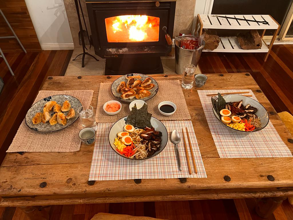 A surprise dinner laid out on a wooden table in front of a lit fireplace: ramen bowls, dumplings and fried dishes.