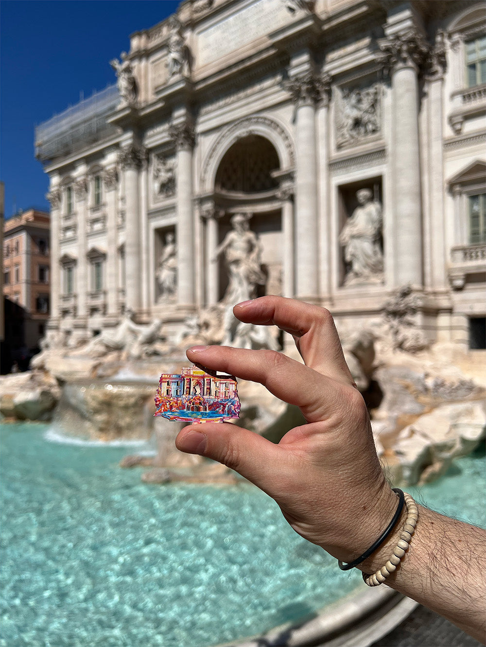 A hand holding a small piece of Flipsi artwork up in front of the Trevi Fountain in Rome.
